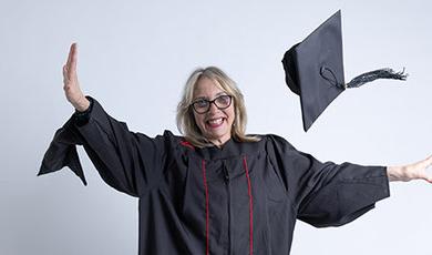 Lesly Freed, wearing her black graduation gown, tosses her cap in the air in front of a white background.