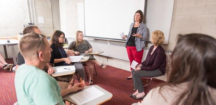 A professor speaking in front of a screen surrounded by a circle of students in desks listening